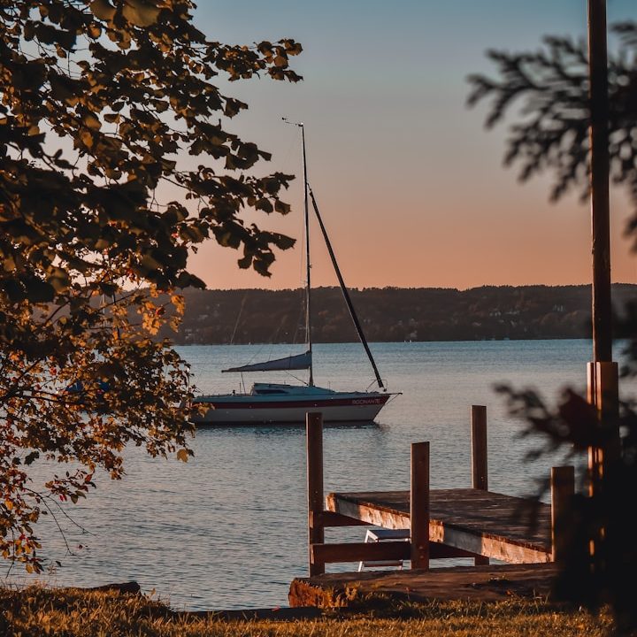 Segelboot auf einem ruhigen See bei Sonnenuntergang, umgeben von Bäumen.